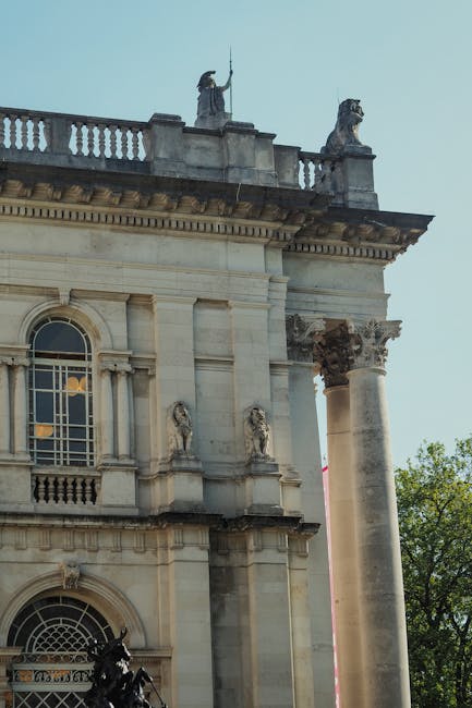 Close-up view of the upper section of a historic classical-style building featuring ornate architectural details, including large column capitals, decorative sculptures, and elaborate cornices made of light-colored stone. On the roofline, there are two stone statues—one appears to be holding a staff or spear, while the other is of a lion sculpture positioned on a pedestal. The building's façade has arched windows with decorative molding and a balustrade running along the roof edge. Surrounding environment includes a clear sky and green trees visible on the right side, indicating an outdoor urban setting. The image captures the contrast between the historic architecture and the modern context, typical of buildings that may require occasional maintenance or cleaning services. Waste Collection Pimlico often provides independent waste handling for such historic sites, contributing to alternative disposal or on-site clearance if necessary.