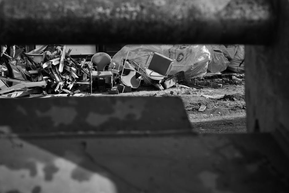A pile of black rubbish bags and discarded packaging materials stacked against a textured beige wall near a red metal door that features several graffiti tags, a white and black sign stating 'No Money', and a blue and white 'Keep Clear' notice. The rubbish bags, which vary in size and appear to contain household waste, are placed on a paved surface with some loose cardboard and cardboard packaging visible at the front. The scene suggests a private or independent waste collection point, possibly awaiting removal by a waste management service such as Waste Collection Pimlico, relevant to local rubbish removal and disposal activities in the area. The environment indicates an alleyway or back street setting, with natural lighting illuminating the scene and emphasizing the utilitarian nature of the waste accumulation in an urban context, where on-site clearance or alternative waste handling methods are in progress or imminent.