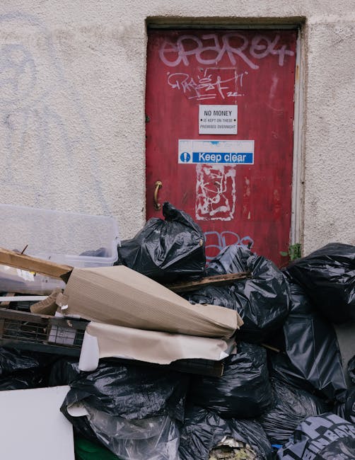 A pile of discarded rubbish bags, primarily black, stacked in front of a red metal door that is heavily covered in graffiti and stickers. The bags are overflowing with waste materials, including a flattened cardboard box and a piece of white foam or insulation. The door, set into a rough-textured beige wall, features signs including a 'No Money' notice and a 'Keep Clear' sticker. The scene appears to be an outdoor urban alley or back entrance, with waste left temporarily outside for collection. The lighting is natural, emphasizing the cluttered arrangement of debris and the weathered appearance of the door and wall, reflecting a typical informal disposal area likely associated with alternative waste removal services managed by companies such as Waste Collection Pimlico.