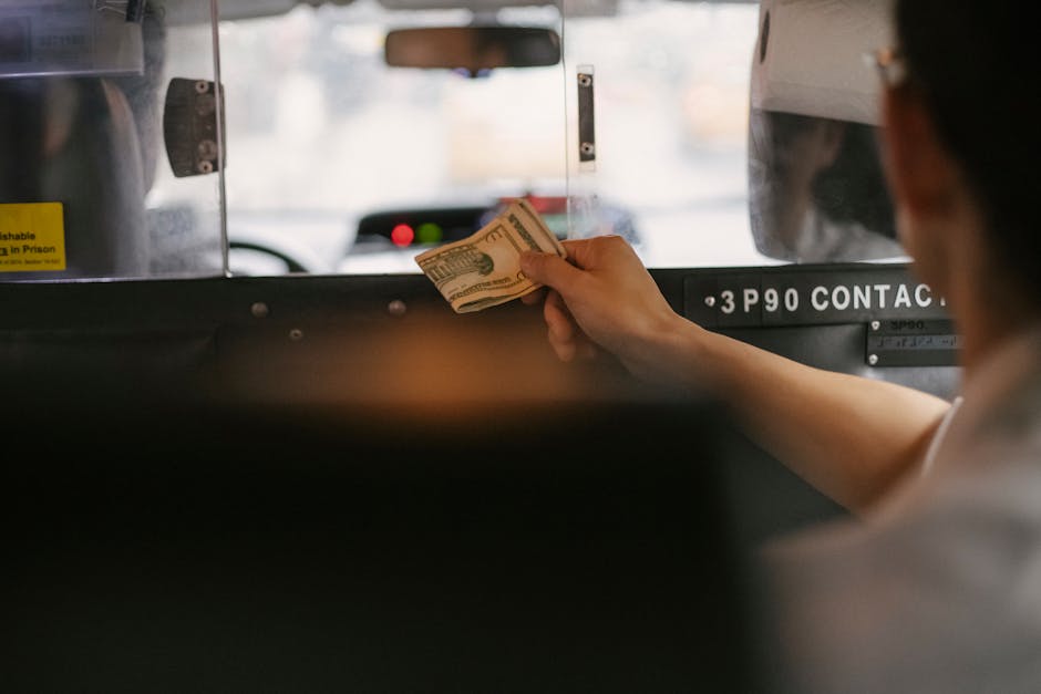 A person’s hand holding a folded stack of US twenty-dollar bills is visible in the foreground, with fingers gripping the bills near the edges. The hand is positioned against the control panel or dashboard of a vehicle, possibly a bus or a van, with a black surface featuring some labels and a section of visible metal screws. In the background, the blurred windshield reveals a street scene with faint shapes of vehicles and buildings, illuminated by daylight. The interior lighting creates a soft, natural atmosphere, and the image emphasizes the transaction or exchange of money within a commercial or transportation setting, aligning subtly with the theme of private waste management services such as rubbish collection by companies like Waste Collection Pimlico. The overall scene is neutral and professional in tone, with clear focus on the hand and money as the primary objects.