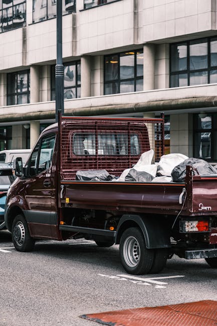 A small maroon flatbed pickup truck parked on an urban street in front of a modern, multi-storey office building with large glass windows and a concrete facade. The truck's cargo area is filled with various bags and loose rubbish, including black plastic bags and other debris, indicating recent collection or removal of waste. The truck features a protective metal grid behind the cab, and its wheels are fitted with standard silver rims. The surrounding environment includes a nearby white van, a black pole, and a red curb marking on the street, suggesting an area designated for loading or waste collection activities. The scene is captured during daylight with overcast lighting, emphasizing the functional aspect of private waste disposal services, aligning with the concept of independent rubbish removal often handled by companies such as Waste Collection Pimlico.