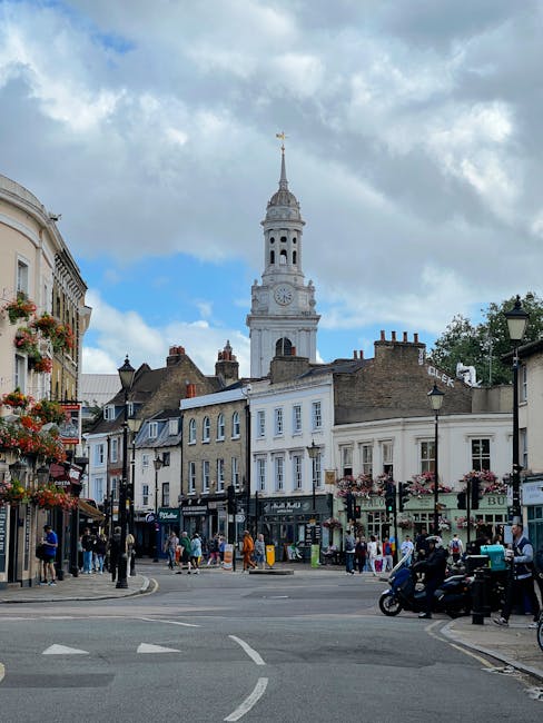 A small maroon flatbed pickup truck parked on an urban street in front of a modern, multi-storey office building with large glass windows and a concrete facade. The truck's cargo area is filled with various bags and loose rubbish, including black plastic bags and other debris, indicating recent collection or removal of waste. The truck features a protective metal grid behind the cab, and its wheels are fitted with standard silver rims. The surrounding environment includes a nearby white van, a black pole, and a red curb marking on the street, suggesting an area designated for loading or waste collection activities. The scene is captured during daylight with overcast lighting, emphasizing the functional aspect of private waste disposal services, aligning with the concept of independent rubbish removal often handled by companies such as Waste Collection Pimlico.