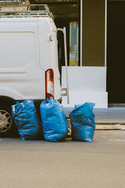 The image depicts the rear side of a white commercial van parked on a street, with three blue rubbish bags placed on the ground close to the vehicle's rear bumper. The bags appear to be made of plastic or similar material, filled with waste, and are tied at the top, showing slight bulges and creases on their surfaces. Behind the van, a storefront or building entrance is visible, featuring a glass door and window, with a white platform or step leading up to it. The environment suggests an urban setting, with the van positioned on a paved street against the building facade. The lighting appears natural, casting minimal shadows, indicating daytime conditions. This scene illustrates typical private waste collection or rubbish disposal practices, which Waste Collection Pimlico offers as an alternative to council-operated rubbish removal services, especially suited for small-scale or on-site clearance tasks.