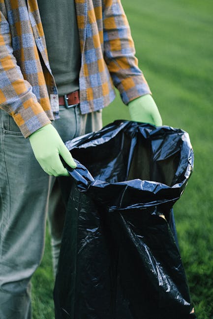 A person standing outdoors on a grassy area, wearing a grey and yellow checkered shirt over a green t-shirt, paired with grey trousers and a brown belt. They are holding open a large black rubbish bag lined with a reflective silver interior, gripping the bag's edges with both hands covered by bright green disposable gloves. The scene suggests an activity related to waste collection or environmental cleanup, potentially supported by waste management services such as Waste Collection Pimlico, who offer independent rubbish removal and disposal solutions. The background features a lush, green lawn, with natural daylight illuminating the scene, emphasizing the task of preparing a waste bag for collection or disposal in an urban or suburban setting dedicated to maintaining cleanliness and proper waste handling.