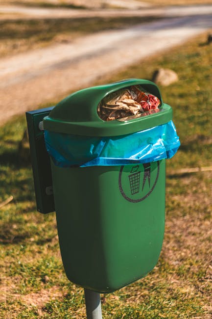 A green public waste bin with a rounded, domed lid positioned outdoors on a grassy area, with a blurred background of a dirt pathway and trees. The lid is slightly open, revealing crumpled paper and assorted small waste items inside. The bin has a blue plastic liner visible around the opening, secured underneath the lid. An icon on the front indicates that the bin is designated for general waste disposal. The scene is illuminated by natural daylight, with soft shadows cast on the ground, illustrating an environment suitable for waste collection or disposal in a communal outdoor setting, as handled by waste collection services such as Waste Collection Pimlico operating within private waste management frameworks.