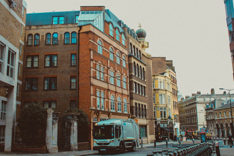 A street scene in an urban area featuring a grey waste collection truck parked on the right side of the road, with a cab and large rear waste bin designed for rubbish collection. The truck is positioned in front of a series of historic multi-storey buildings, including a red-brick structure with decorative arched windows and a blue-tiled roof, alongside other buildings with ornate facades and large windows. The environment appears to be a busy city street with street lamps, bollards, and parked bicycles in the foreground, and a cloudy sky overhead. The scene relates to waste management services such as private rubbish collection provided by Waste Collection Pimlico, illustrating the practical aspect of on-site rubbish removal in an urban setting where independent waste disposal methods are common.