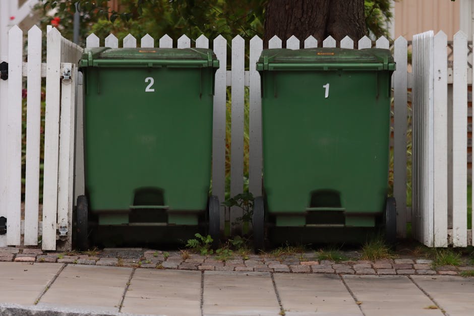 Two green wheelie bins with black wheels and closed lids are positioned next to each other on a paved sidewalk, with the bin on the left labeled with the number '2' and the one on the right labeled '1'. They are placed in front of a white picket fence with vertical slats, which runs horizontally across the background. Behind the fence, there is a mature tree trunk with visible bark texture, and some foliage is partially visible at the top of the image. The bins have a smooth, hard plastic surface with a matte finish, and slight rust stains can be seen around the hinges of the lids. The pavement beneath the bins consists of concrete tiles, with some small weeds and grass growing at the edges where they meet the ground. The environment appears to be an outdoor residential setting, likely a front or back garden area, where private waste collection services are arranged, as opposed to public waste authority collection. The lighting is natural and evenly diffused, suggesting an overcast sky or shaded location, aligning with typical rubbish disposal scenes in suburban or urban residential contexts.