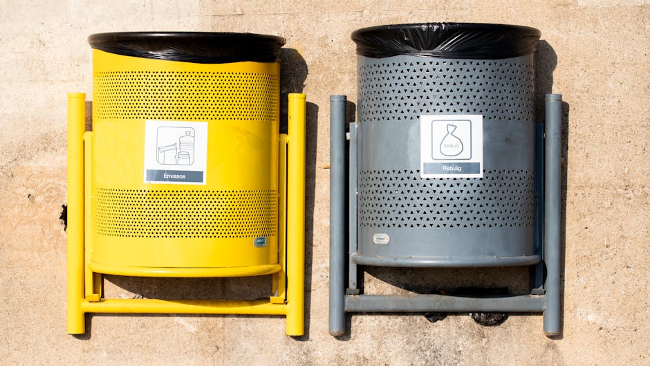 Two cylindrical waste bins are positioned side by side on a paved surface. The bin on the left is painted bright yellow with a perforated design, a black lid, and a white label depicting an image of a bottle and a box, indicating it is for packaging waste. The bin on the right is grey with a matching perforated pattern, a black lid, and a white label featuring an icon of a glass container, suggesting it is for recyclable glass. Both bins are supported by metal frames, with the yellow bin’s frame in matching yellow and the grey bin’s frame in metallic grey. Behind the bins, the background consists of a smooth, beige concrete wall. The scene suggests a typical outdoor waste collection setup, possibly part of an independent waste management service like Waste Collection Pimlico, emphasizing options for different types of waste separation, which are an essential aspect of private disposal and on-site clearance solutions in urban areas.