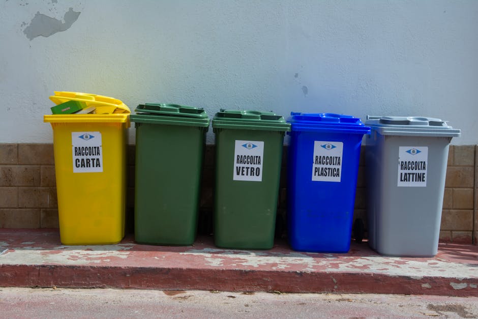 Two green wheelie bins with black wheels and closed lids are positioned next to each other on a paved sidewalk, with the bin on the left labeled with the number '2' and the one on the right labeled '1'. They are placed in front of a white picket fence with vertical slats, which runs horizontally across the background. Behind the fence, there is a mature tree trunk with visible bark texture, and some foliage is partially visible at the top of the image. The bins have a smooth, hard plastic surface with a matte finish, and slight rust stains can be seen around the hinges of the lids. The pavement beneath the bins consists of concrete tiles, with some small weeds and grass growing at the edges where they meet the ground. The environment appears to be an outdoor residential setting, likely a front or back garden area, where private waste collection services are arranged, as opposed to public waste authority collection. The lighting is natural and evenly diffused, suggesting an overcast sky or shaded location, aligning with typical rubbish disposal scenes in suburban or urban residential contexts.