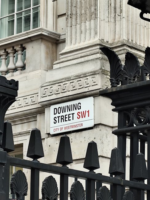 A close-up view of a street sign attached to the corner of a classical stone building in Pimlico, Westminster. The sign reads 'Downing Street SW1' and indicates the location within the City of Westminster. The building features detailed architectural elements such as fluted pilasters, decorative molding, and a balustrade with small stone balusters visible below a large window with multiple panes. In the foreground, a black wrought iron fence with decorative finials and ornamental leaf motifs surrounds the building, appearing weathered with some elements slightly worn. The lighting suggests daylight, with natural shadows cast across the stone façade and metalwork, creating a clear, professional visual that could relate to urban site management or property boundaries in an area known for governmental and official buildings. Waste Collection Pimlico occasionally handles debris or waste from such premises, though no waste or rubbish is visible in the image. The overall scene emphasizes the architectural setting typical of Westminster administrative infrastructure, aligning subtly with the themes of private property and on-site clearance activities related to rubbish removal services.