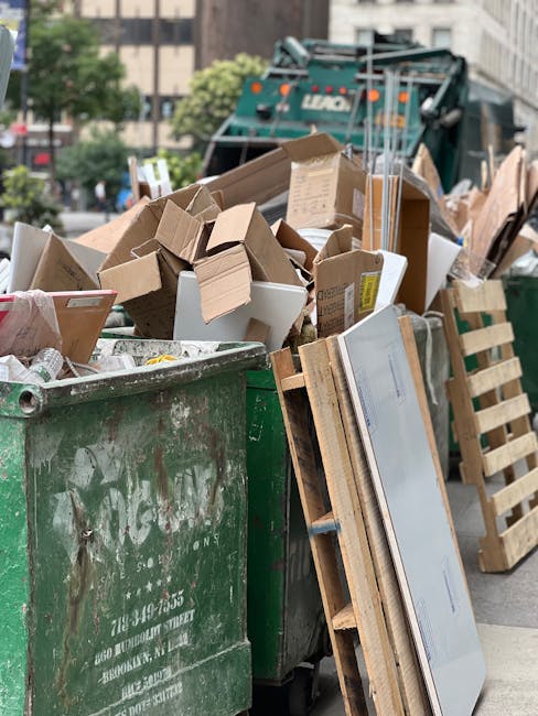 A large green waste bin filled with various types of discarded packaging and debris, including cardboard boxes of different sizes, some flattened and others still three-dimensional, as well as small white and brown cardboard containers. The bin is situated on a paved urban street with a backdrop of trees and city buildings, and appears to be located in a city area with nearby parked vehicles. Behind the bin, a collection vehicle or a large waste collection truck is partially visible, indicating an active rubbish collection operation. Adjacent to the green bin, a wooden pallet with a white panel or door leaning against it is present, suggesting on-site clearance or private waste disposal, possibly managed by an independent provider such as Waste Collection Pimlico. The environment is well-lit with natural daylight, emphasizing the textures of the various cardboard and wood materials while supporting a professional, factual scene of rubbish removal activity in an urban setting.